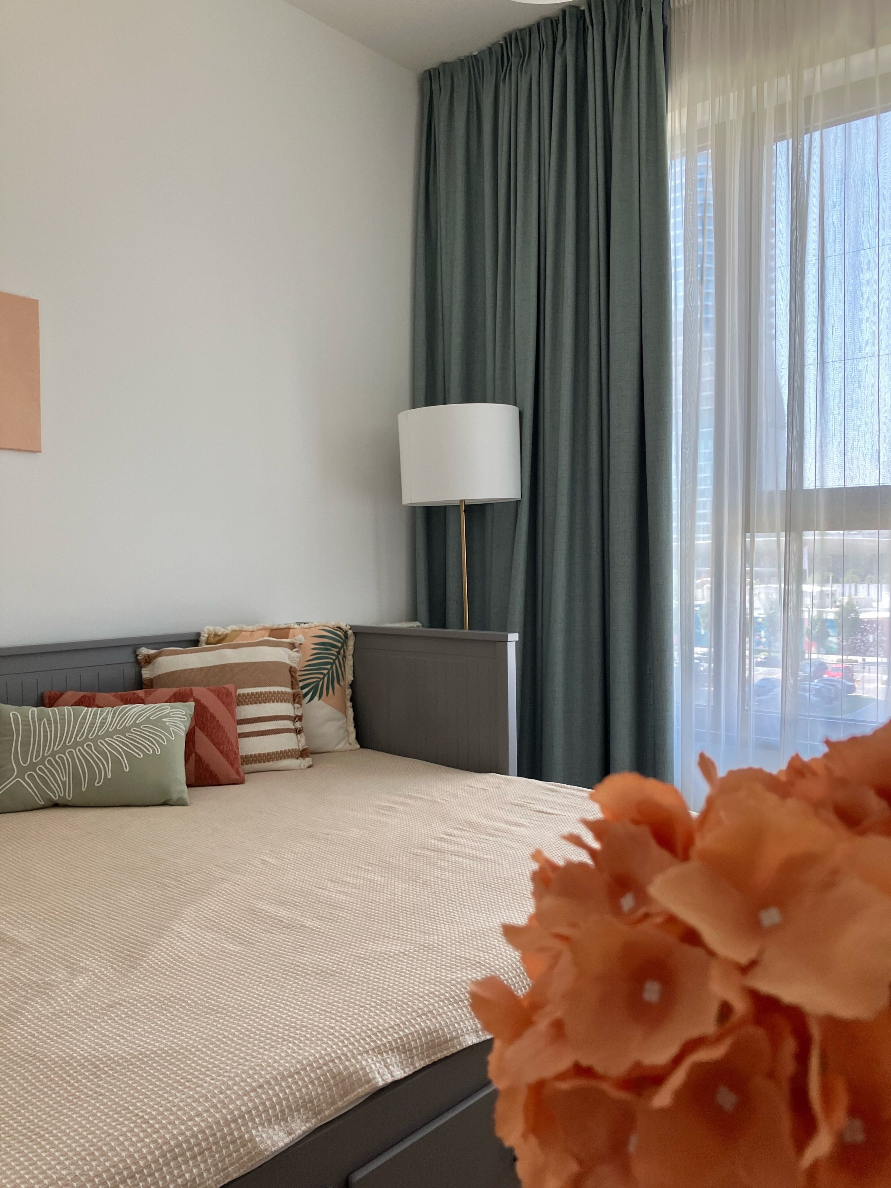 A bedroom with a bed, a nightstand, a lamp, and a view of the outside through sheer curtains.