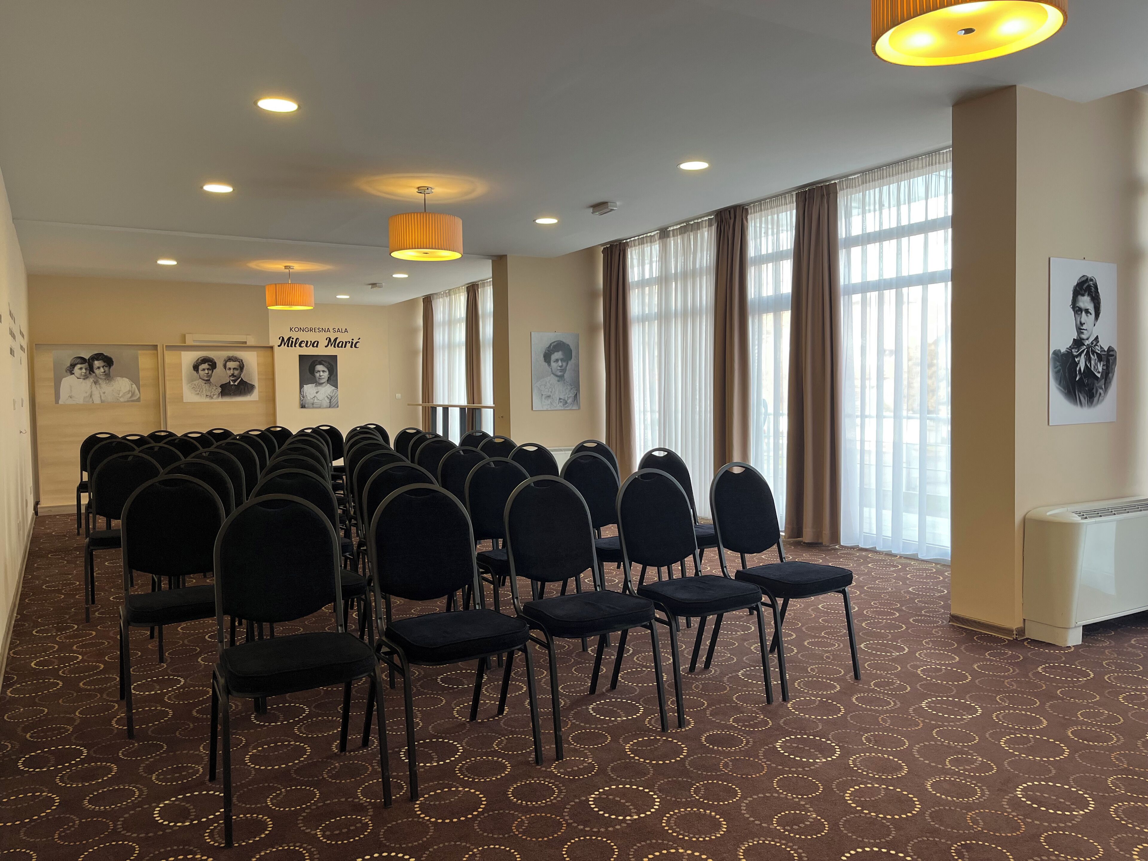 Une salle de conférence avec des rangées de chaises, une moquette à motifs et de grandes fenêtres avec des rideaux.