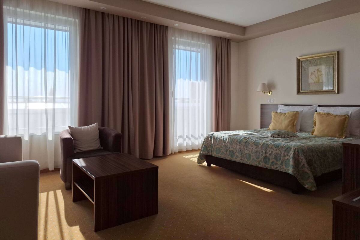 A hotel room with a bed, a desk, a chair, and large windows with curtains.