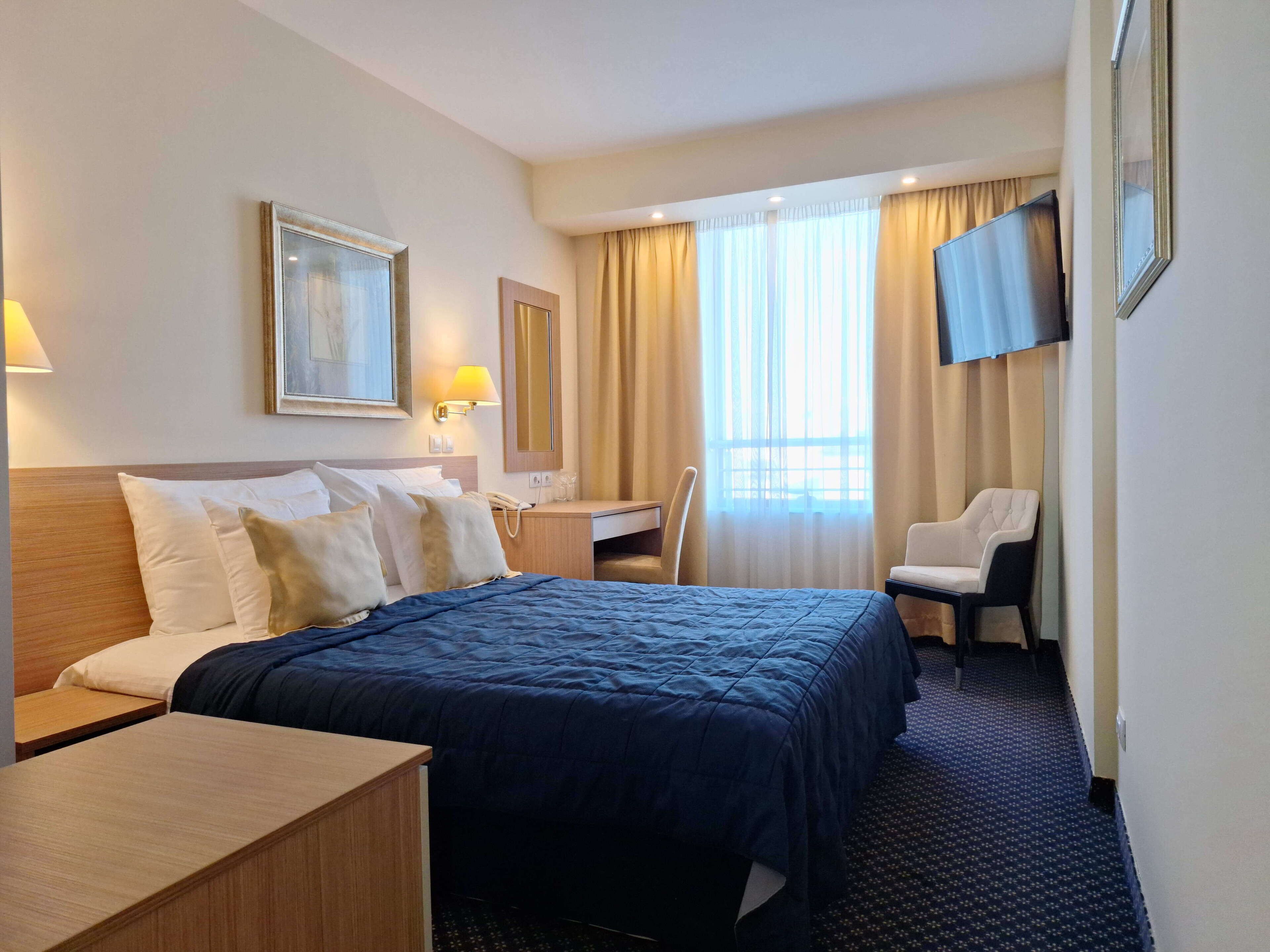 A hotel room with a large bed, a desk, a chair, a TV, and a window with curtains.