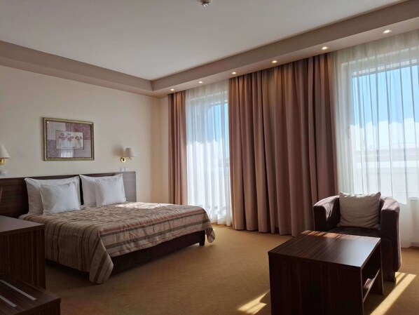A hotel room with a bed, a desk, a chair, and large windows with curtains.