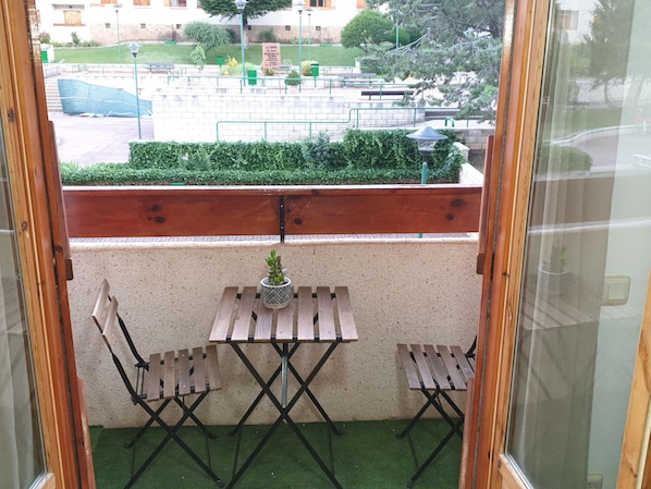 A small balcony with a wooden table and two chairs, a potted plant, and a view of a park and buildings in the background.