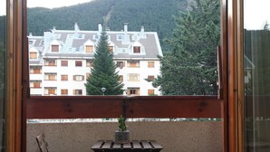 A balcony with two wooden chairs and a small table, overlooking a row of buildings with multiple windows and a mountain in the background.