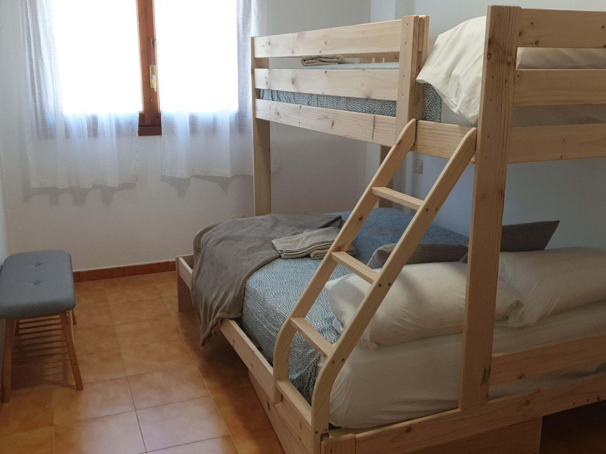 Ein Etagenbett mit einer Leiter, ein kleiner Hocker und ein Fenster mit durchsichtigen Vorhängen.