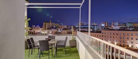 A balcony with a table and chairs, a potted plant, and a view of a cityscape at night.