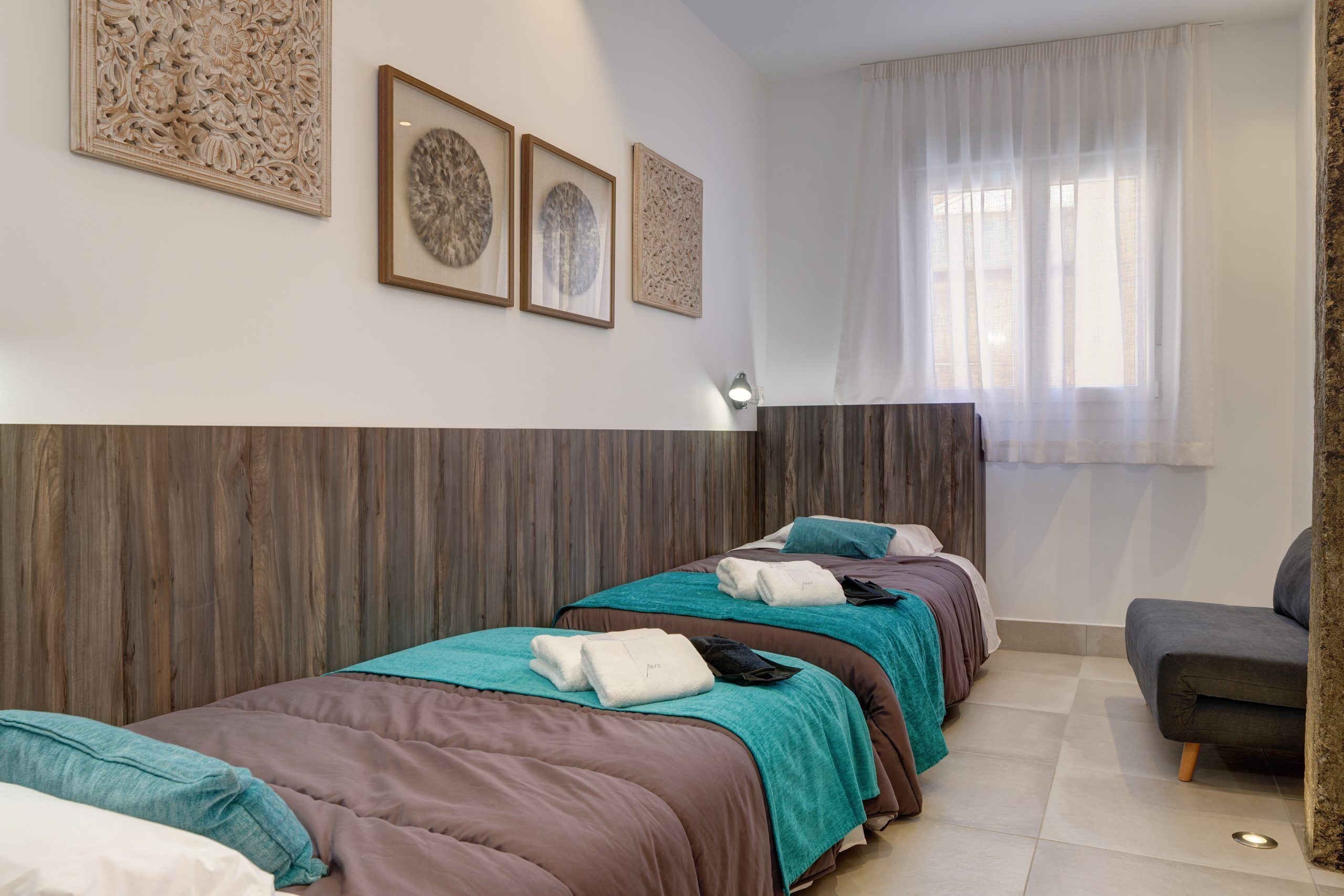 Une chambre d’hôtel avec deux lits, une tête de lit en bois, une fenêtre avec des rideaux légers et des œuvres d’art encadrées accrochées au mur.