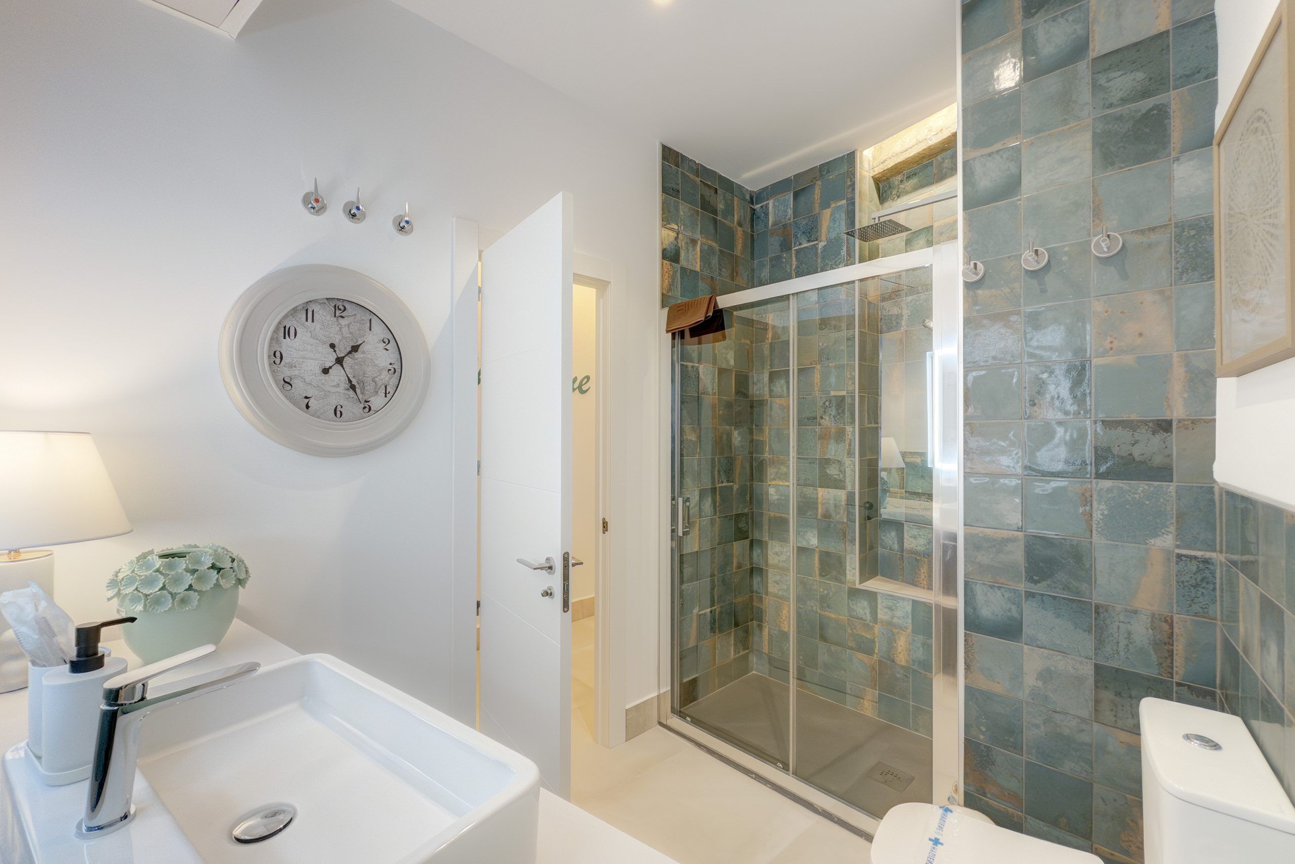 A modern bathroom with a glass shower enclosure, a white sink, and a round wall clock.
