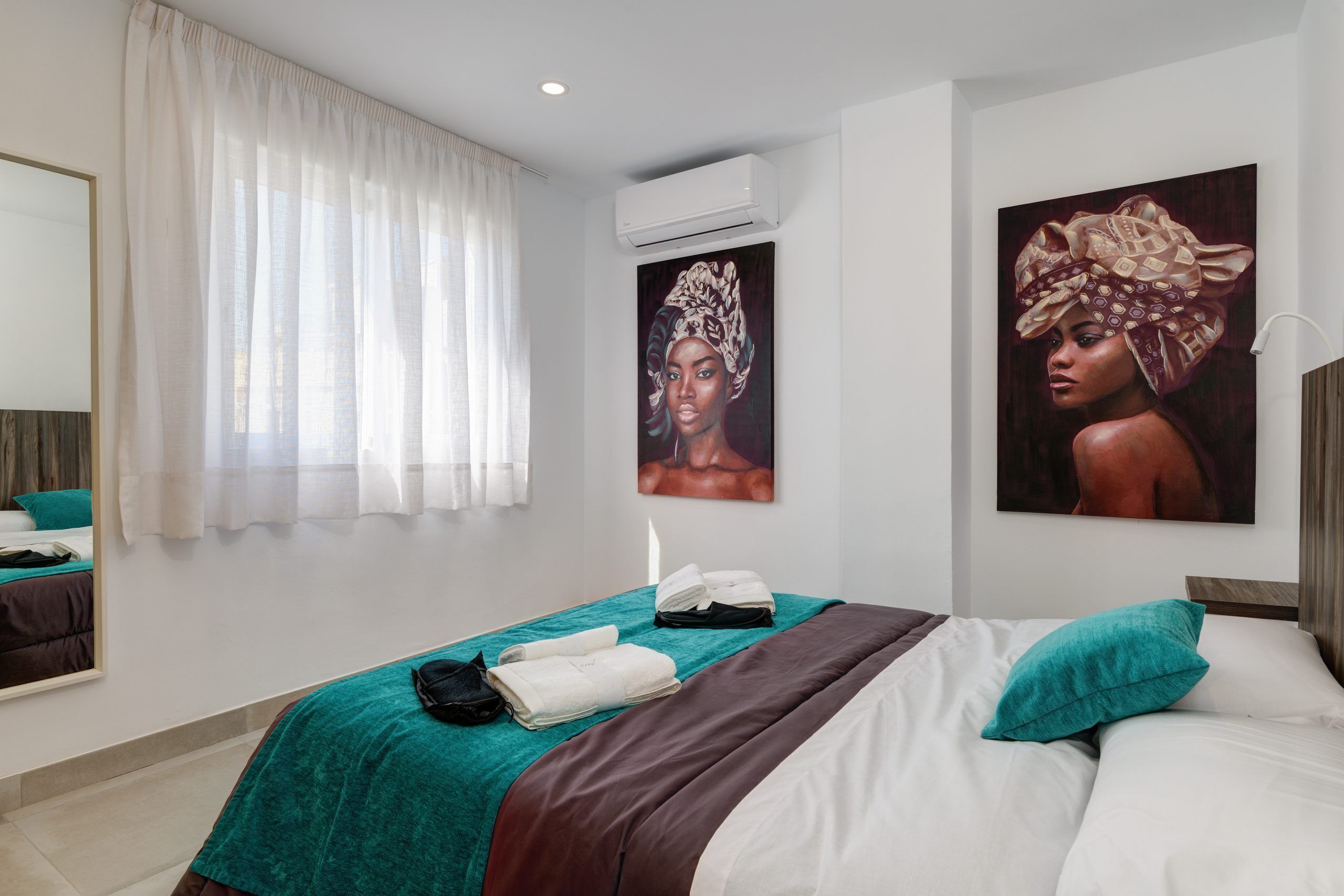 A hotel room with a bed, a mirror, a window with curtains, and artwork on the walls.