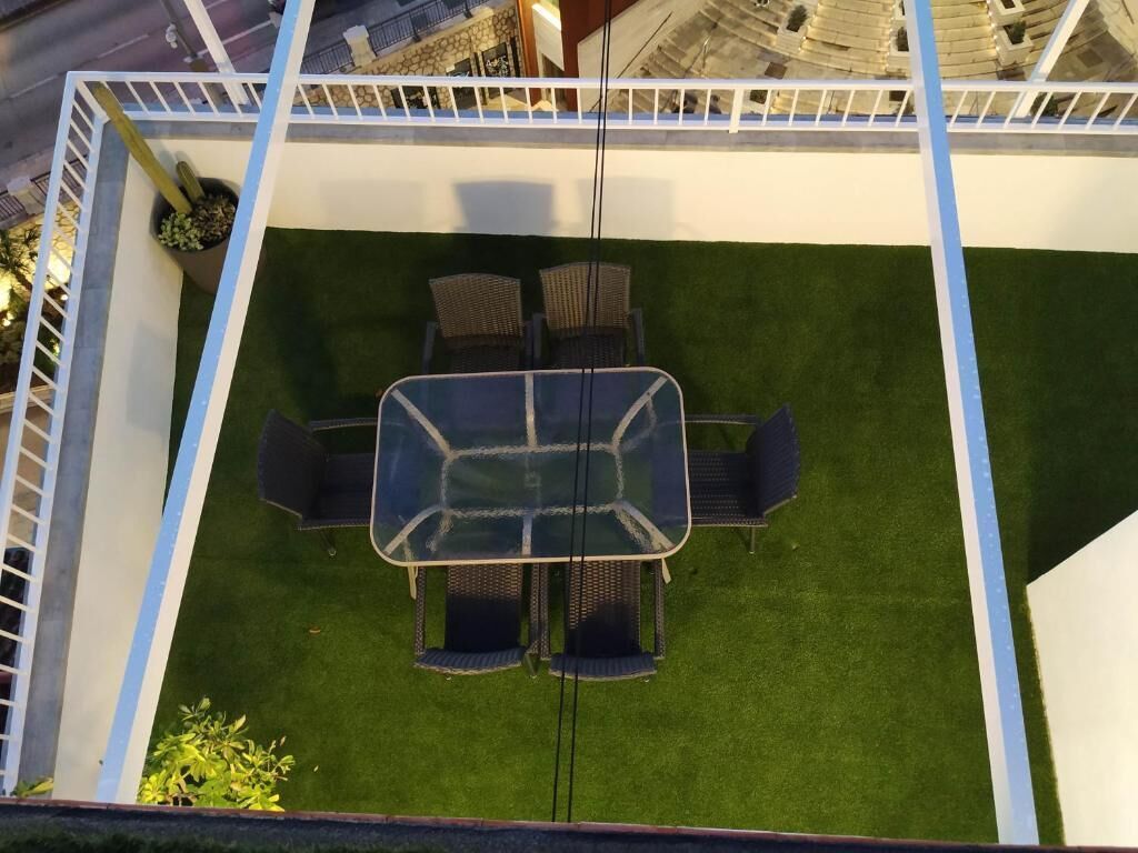An outdoor patio area with a glass-top table and four chairs, surrounded by a white fence and green artificial grass.