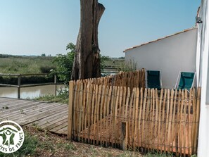 A rustic wooden deck with a bamboo fence, a white building with a red roof, and a view of a river and greenery. - 4-bedroom cottage with wellness and terraces in the heart of Marais Breton (Bois-de-Céné)