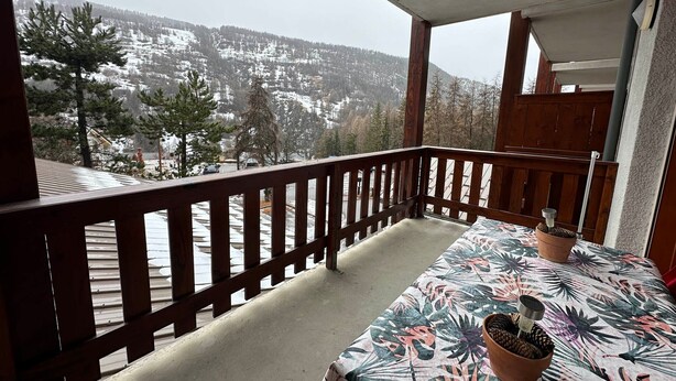 A balcony with a wooden railing, a table with a floral tablecloth, and a view of snow-covered mountains. - Super Duplex in Vars - 2 Bedrooms, Terrace, Parking (Vars)