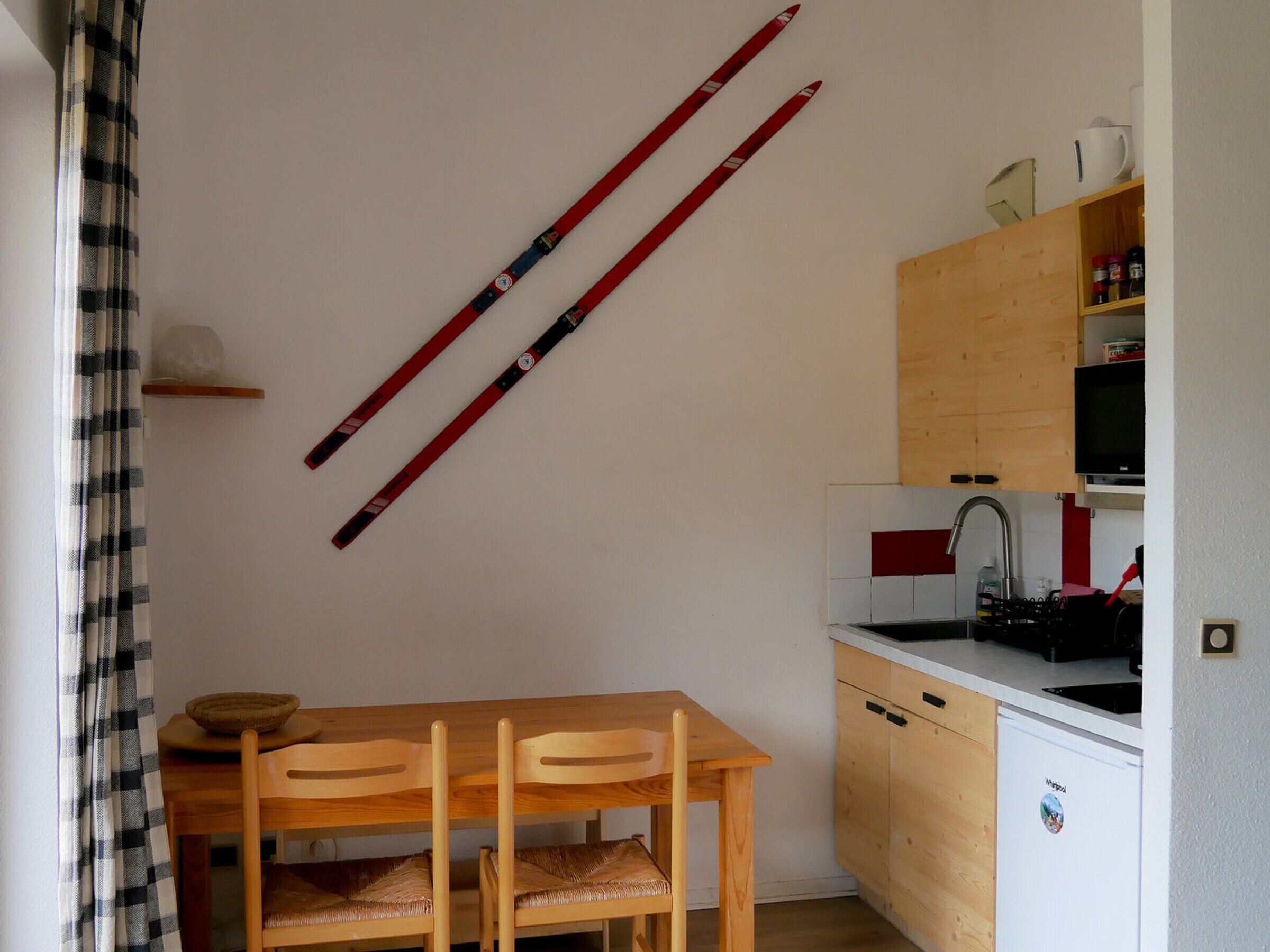 A small kitchen with wooden cabinets, a sink, and a stove. There is a table with two chairs and a pair of skis leaning against the wall.