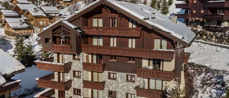 Un albergue de varios pisos con construcción de piedra y madera, con balcones y grandes ventanales, rodeado por terreno cubierto de nieve y otros edificios similares.