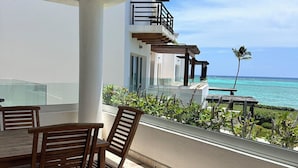 A balcony with a wooden table and chairs, overlooking a turquoise ocean and a white building with a balcony.