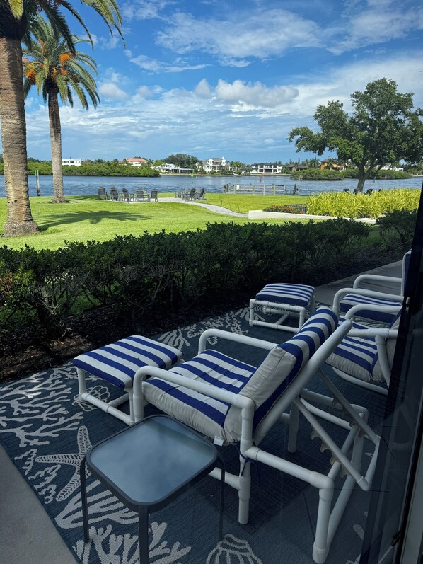 A patio with lounge chairs and a table, overlooking a body of water and a landscaped area with palm trees and a grassy lawn.