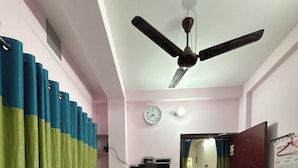 A hotel room with a bed, a ceiling fan, a clock, a TV, and a calendar on the wall. - Green Place Guest House (Bankura)
