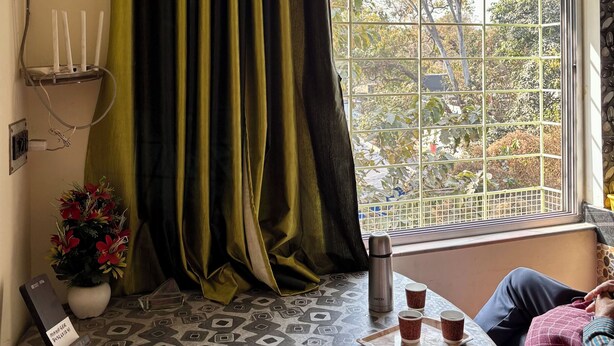 A room with a patterned table, a window with a metal grid, a potted plant, and a red chair. - Green Place Guest House (Bankura)