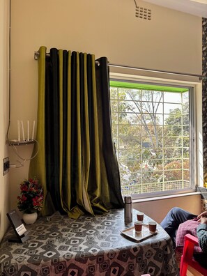 A room with a patterned table, a window with a metal grid, a potted plant, and a red chair. - Green Place Guest House (Bankura)