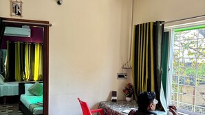 A room with a ceiling fan, a bed, a table with chairs, a window with curtains, and a door leading to another room. - Green Place Guest House (Bankura)