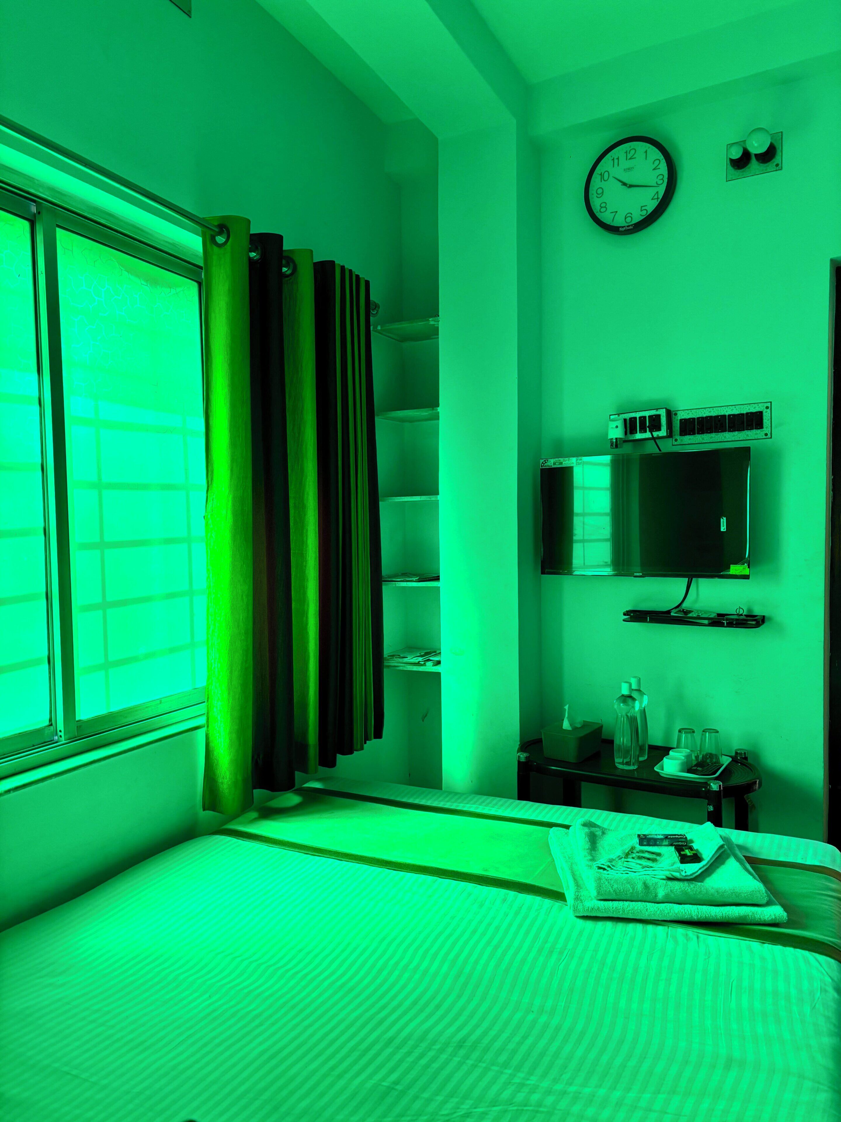 Una habitación con una cama, una ventana con cortinas, un televisor en la pared, una estantería con objetos y un reloj.