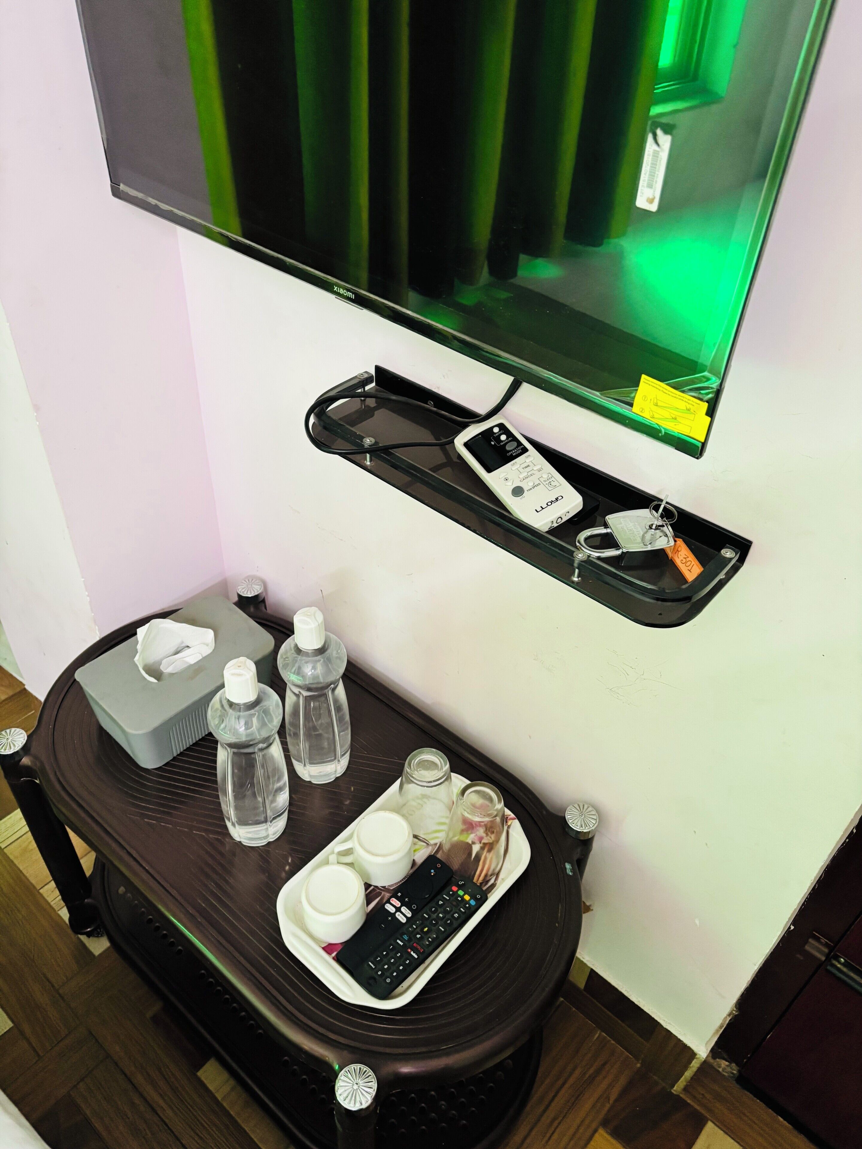 Une chambre d’hôtel équipée d’un téléviseur à écran plat fixé au mur, d’une petite table avec une télécommande, des bouteilles d’eau, une boîte de mouchoirs et un verre d’eau posés sur une petite table.