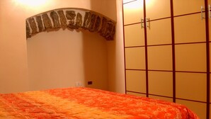 A room with a bed covered in an orange patterned bedspread, a wall-mounted shelf with a decorative item, and a wardrobe with a grid pattern. - Appartamenti nel Centro Storico di Siculiana (Siculiana)