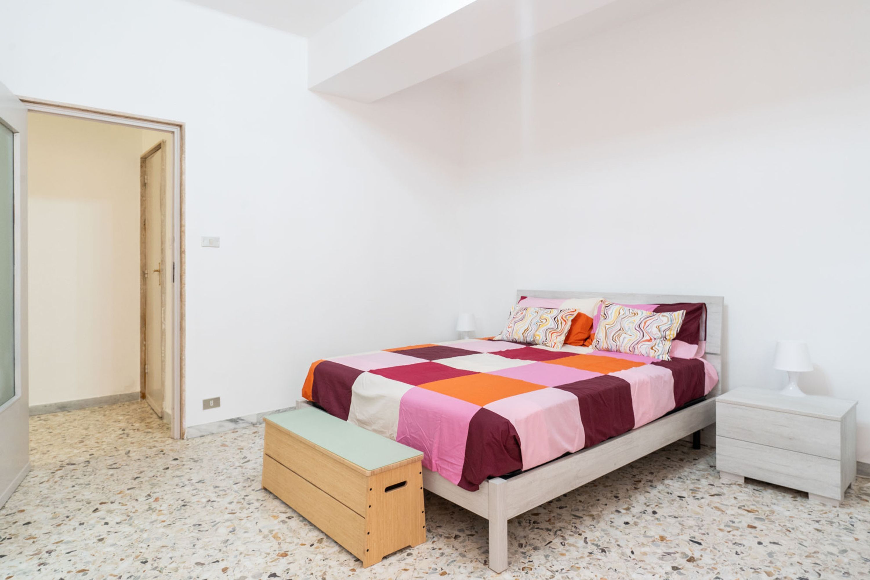A bedroom with a bed, bedside table, bench, and a door.