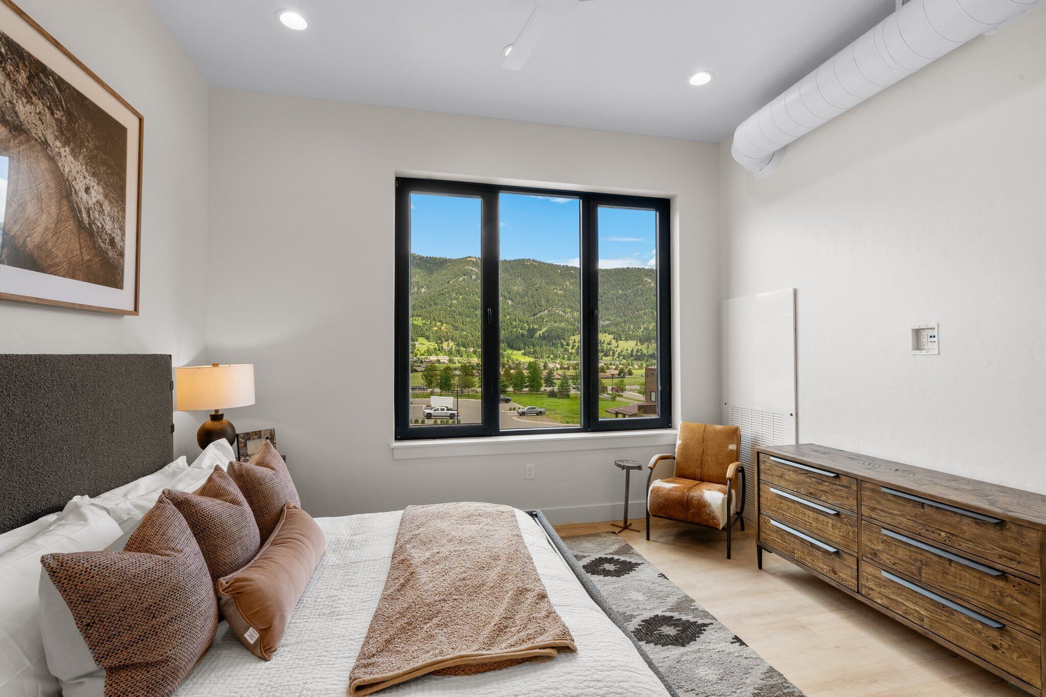 Ein Schlafzimmer mit Bett, Stuhl, Kommode, Fenster und Blick ins Grüne sowie einer Lampe.