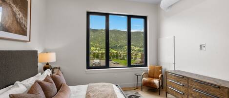 Ein Schlafzimmer mit Bett, Stuhl, Kommode, Fenster und Blick ins Grüne sowie einer Lampe.