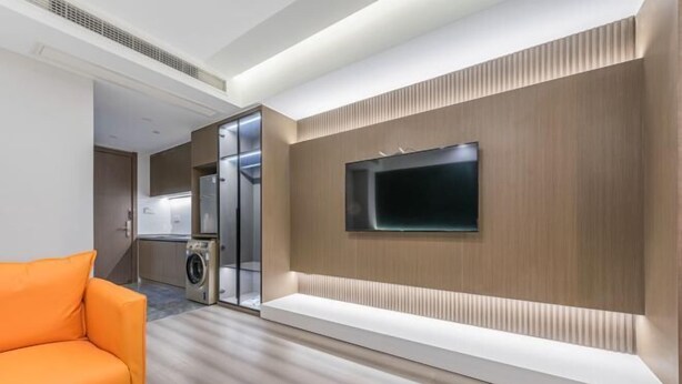 A modern living room with a flat-screen TV mounted on a wooden panel wall, a sofa, and a washing machine in the background. - Ningbo jinfu hotel apartment (Ningbo)