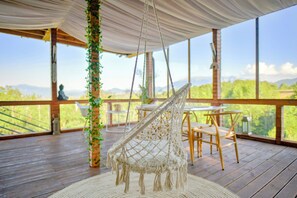 Uma cadeira rede suspensa no teto, um deck de madeira e janelas amplas com vista para o exterior.