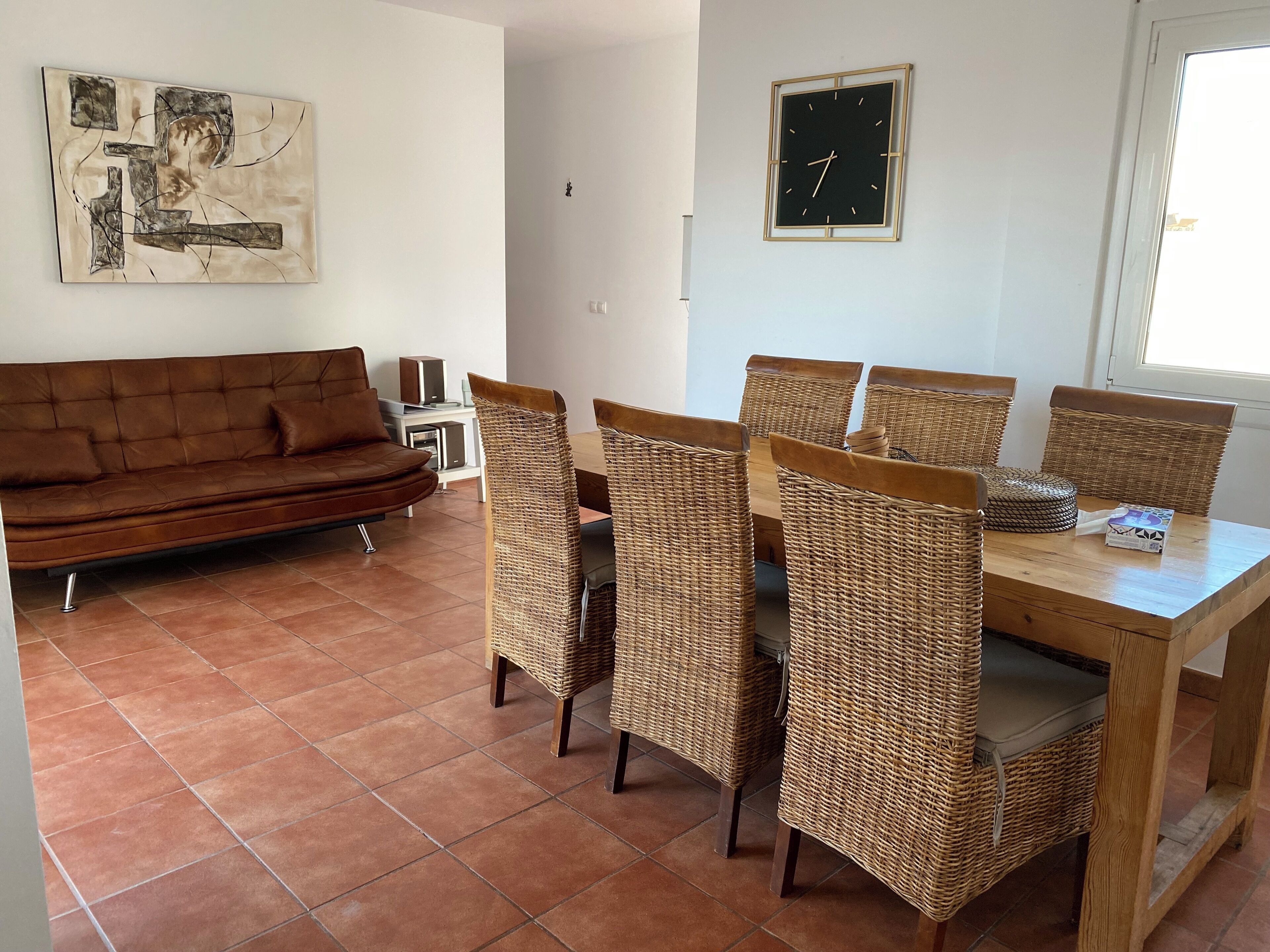 A living room with a brown sofa, wicker chairs, a wooden dining table, and a wall clock.