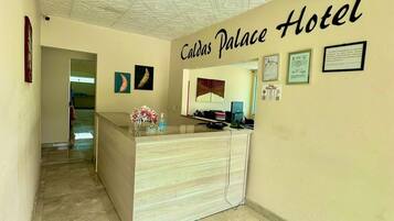 A hotel reception area with a reception desk, a ceiling with a geometric pattern, and a wall with the hotel's name and framed certificates.