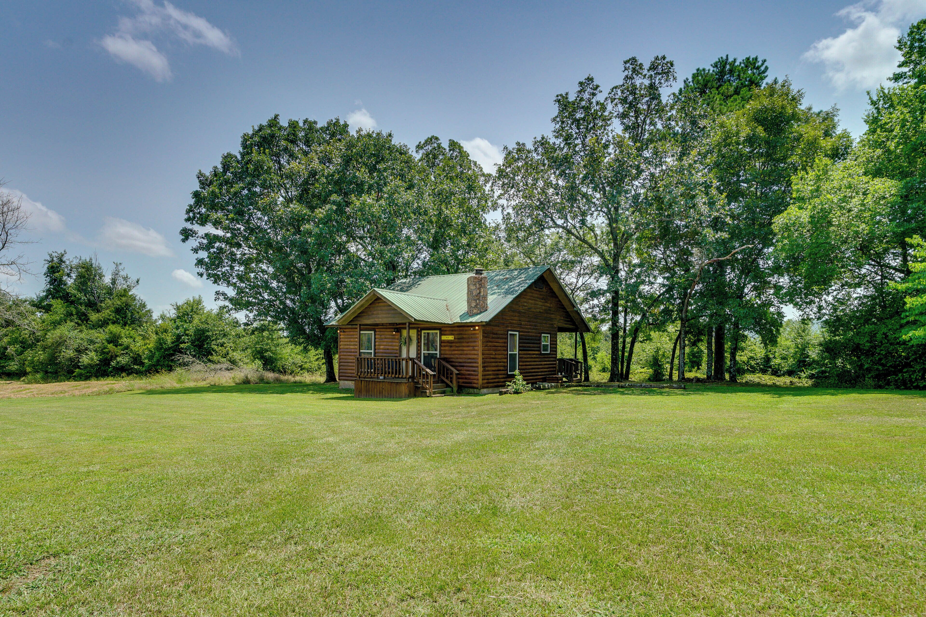 A wooden cabin with a porch and a chimney, surrounded by a grassy field and trees.