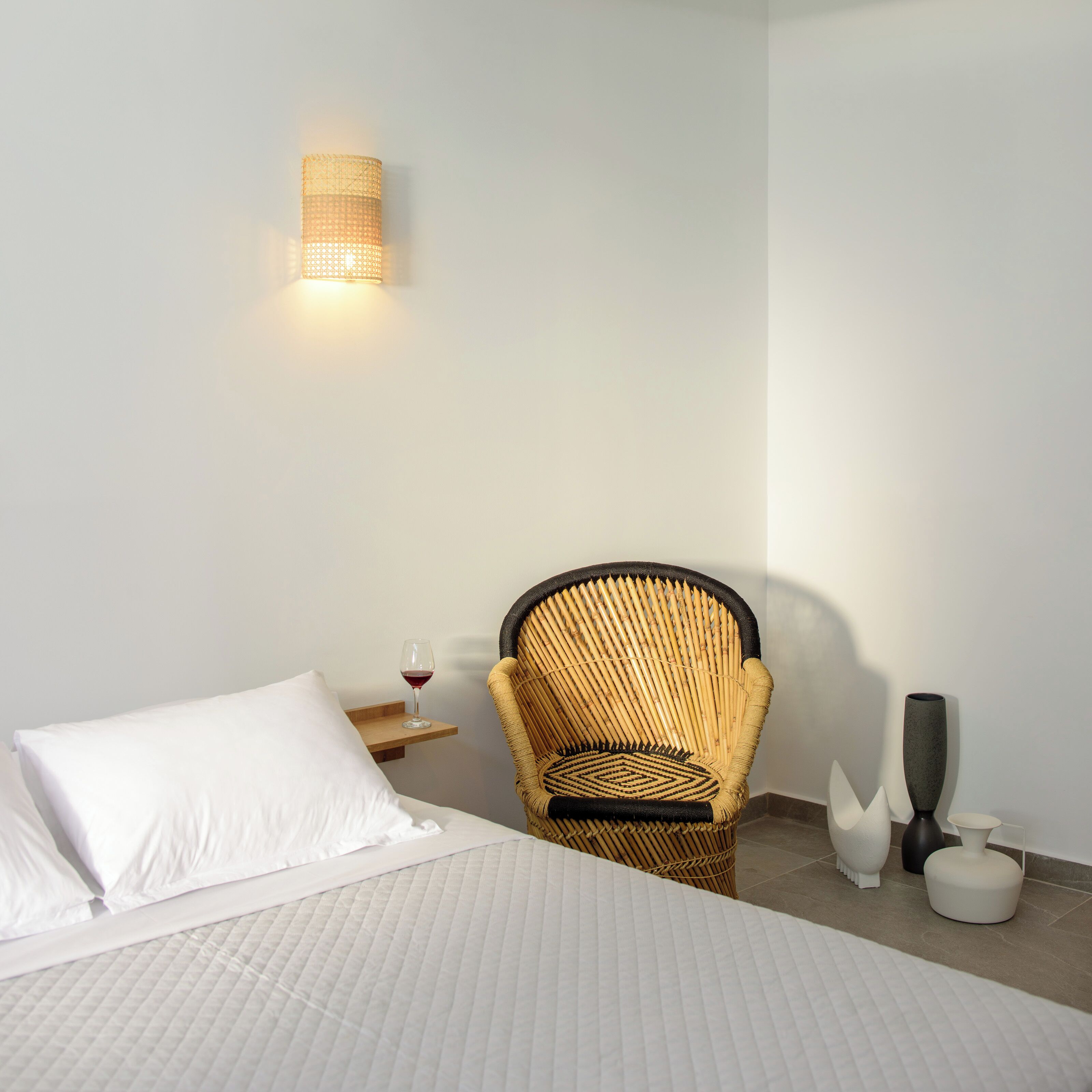 Ein Schlafzimmer mit einem Bett, einem Korbsessel, einem Nachttisch mit einem Weinglas und einer Wandleuchte.