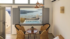 A bedroom with a large bed, two wicker chairs, a small table with a bottle and glasses, and a view of the sea through a window. - Elia Caldera Suites (Santorini)
