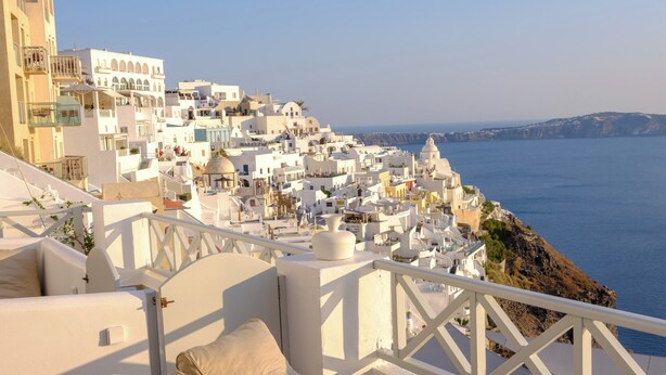 A seaside view with white buildings, a balcony with beige cushions, and a clear blue sky. - Elia Caldera Suites (Santorini)