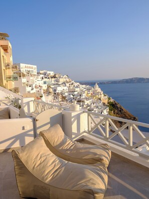 A seaside view with white buildings, a balcony with beige cushions, and a clear blue sky. - Elia Caldera Suites (Santorini)