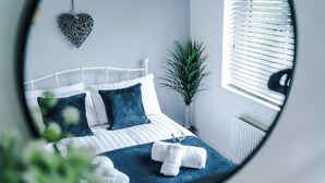 A bedroom with a white metal bed frame, a blue bedspread, and two pillows. There is a potted plant beside the window and a heart-shaped decoration on the wall. - NEW! Stunning Bungalow, Ideal for Groups, Parking! (Wrexham)