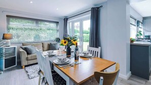 A modern dining area with a wooden table set for four, a sofa, and a kitchen area visible in the background. - NEW! Stunning Bungalow, Ideal for Groups, Parking! (Wrexham)