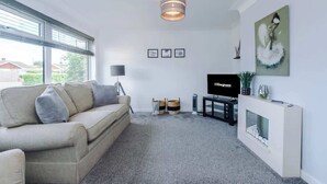 A modern living room with a sofa, a TV, a fireplace, and a large window. - NEW! Stunning Bungalow, Ideal for Groups, Parking! (Wrexham)
