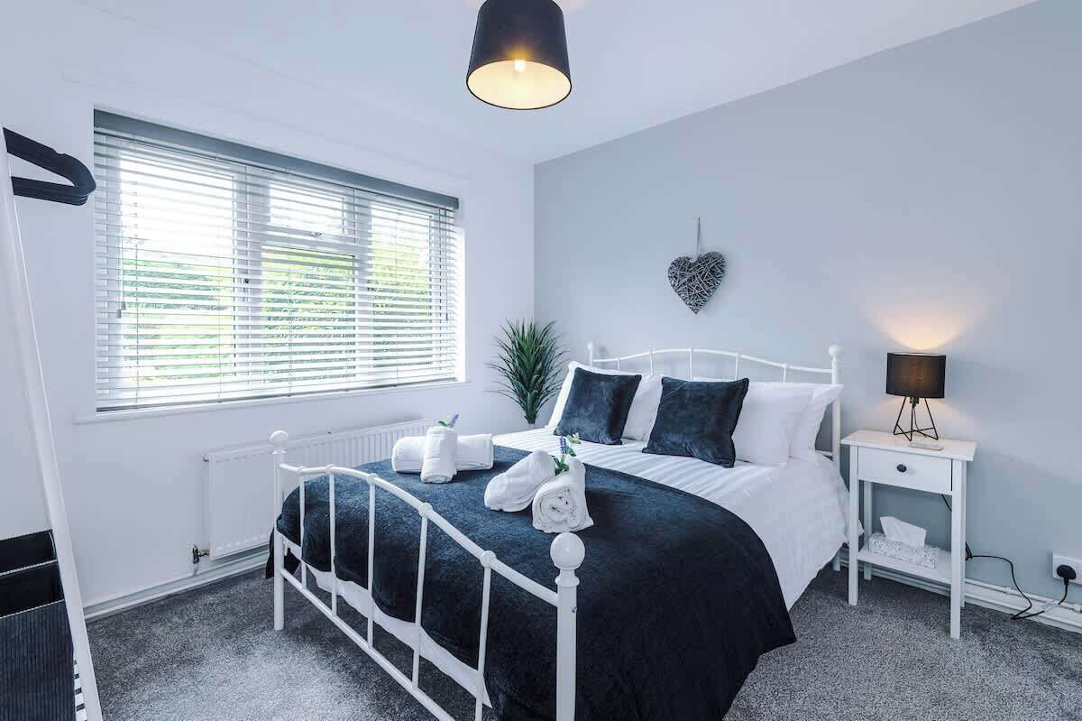 Un dormitorio con estructura de cama de metal blanco, ropa de cama oscura, una lámpara de noche y una decoración con forma de corazón en la pared.