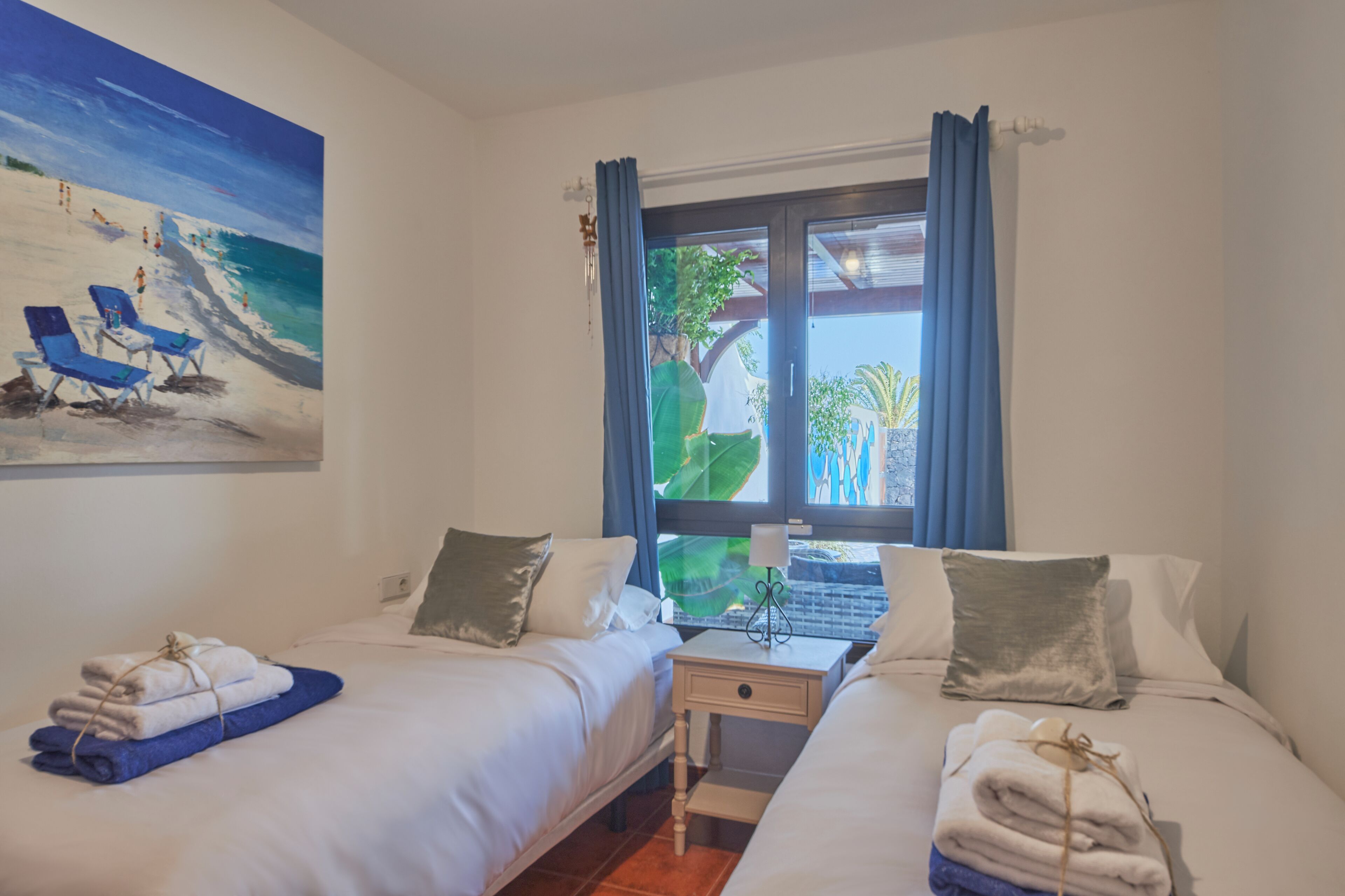 Une chambre d’hôtel avec deux lits, un tableau représentant une scène de plage, une fenêtre avec des rideaux et une vue sur la verdure à l’extérieur.