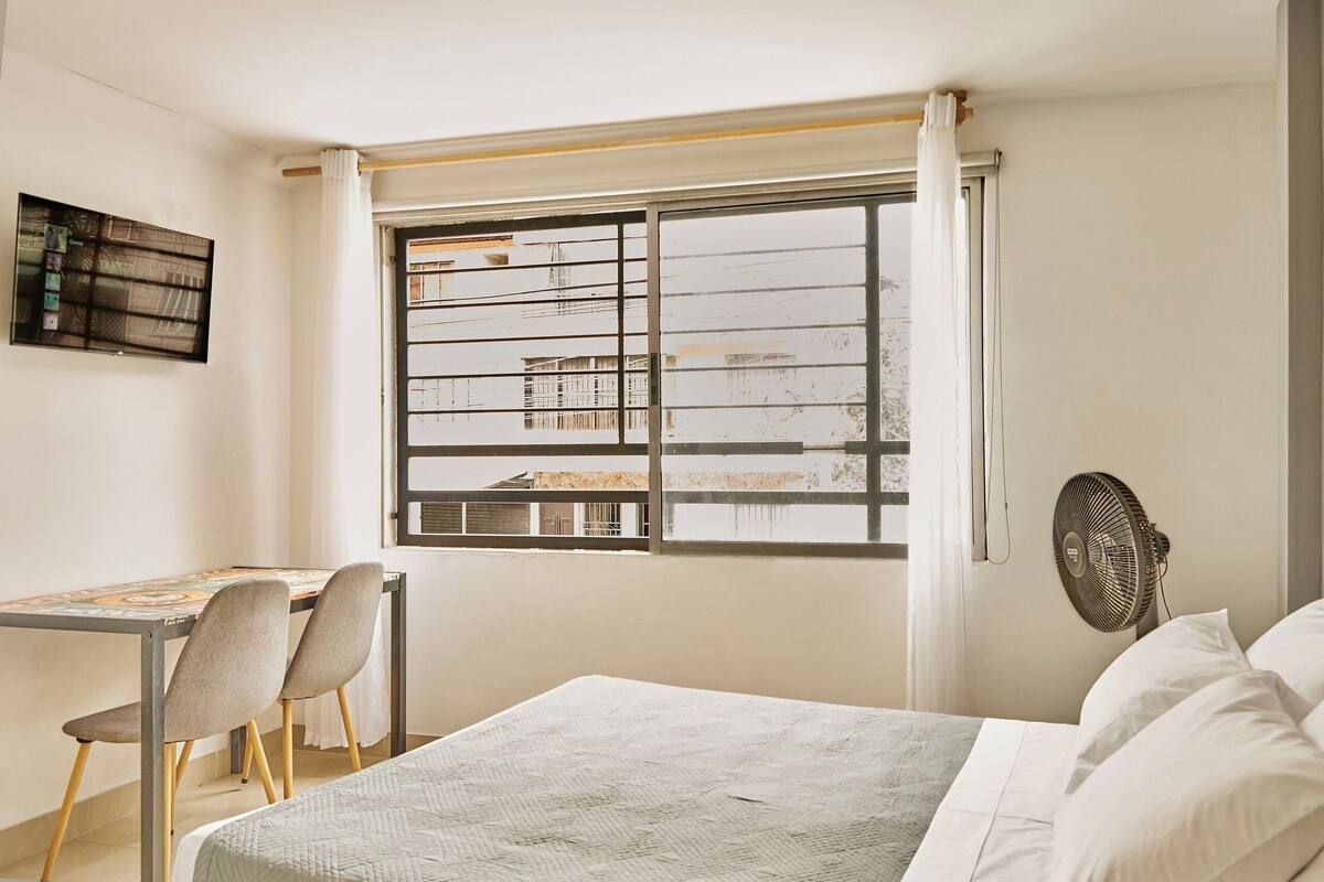 Een slaapkamer met een bed, een bureau met stoelen, een raam met uitzicht op gebouwen en een staande ventilator.