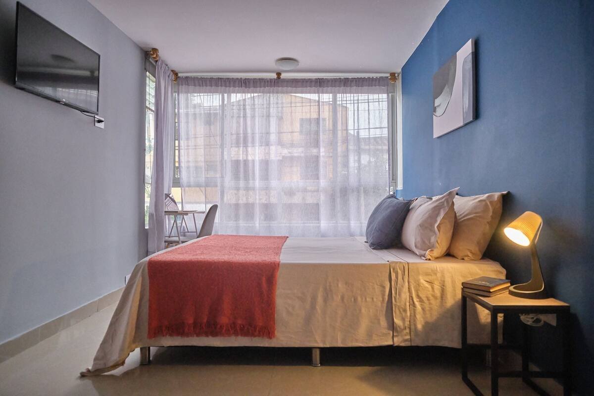 Een slaapkamer met een bed, een nachtkastje met een lamp, een stoel en een raam met dunne gordijnen.