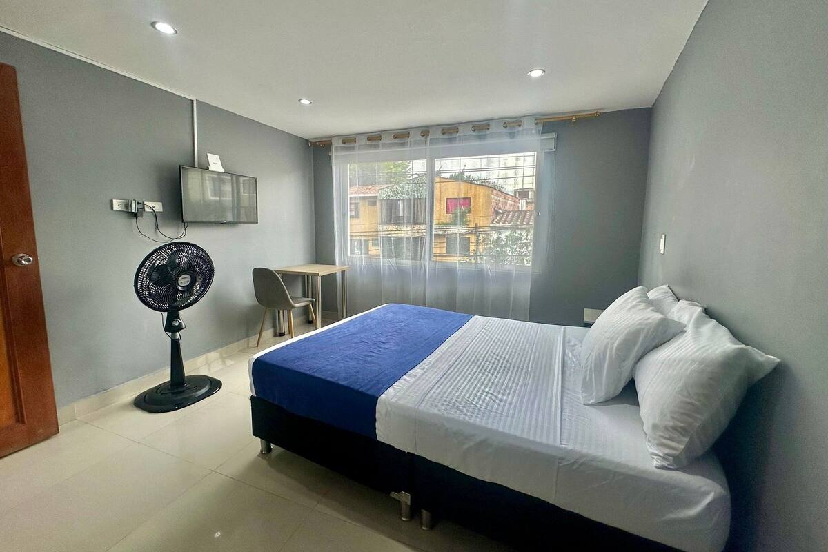 Een slaapkamer met een bed, een bureau, een stoel, een ventilator en een raam met uitzicht op gebouwen.