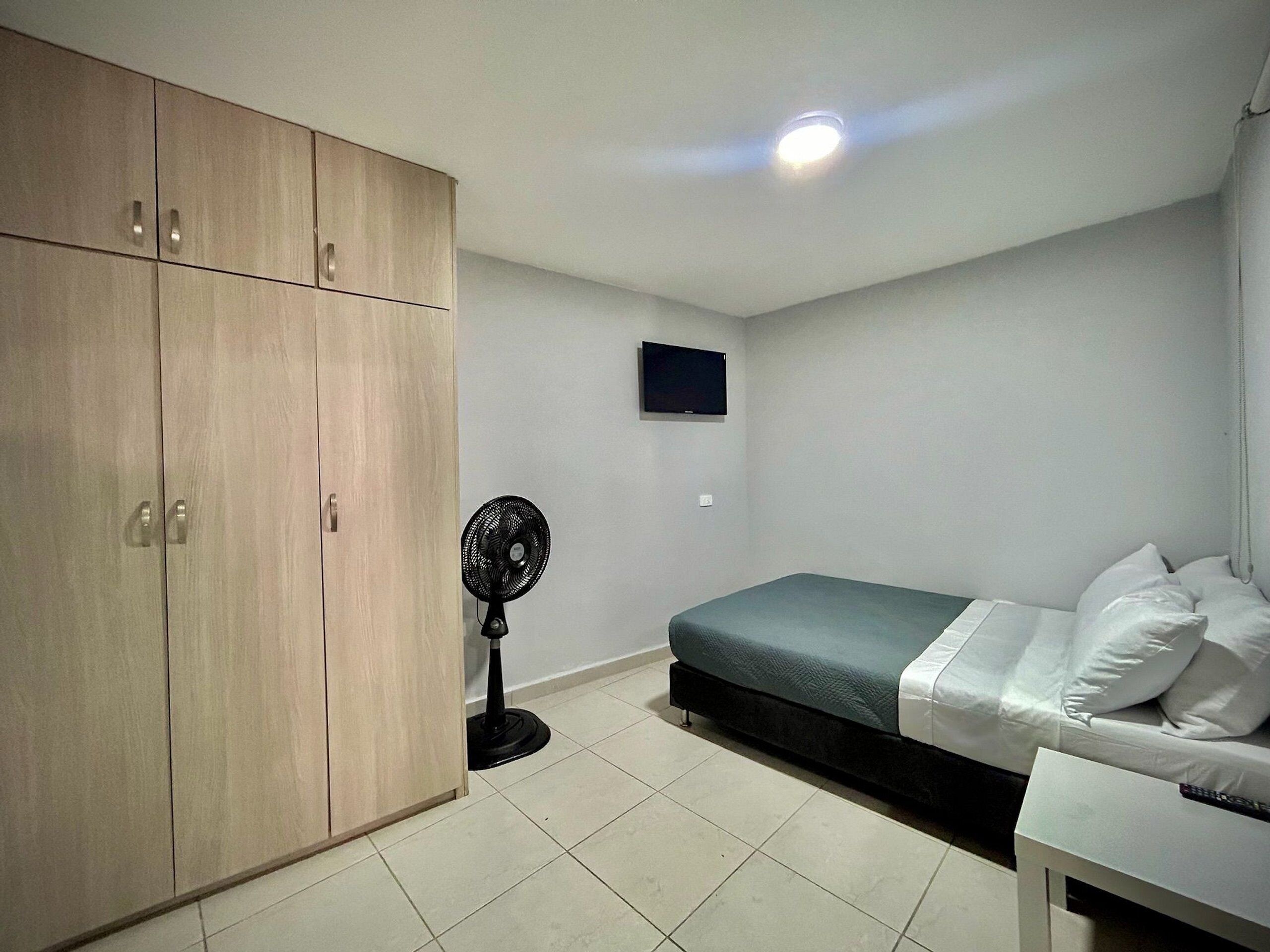 Une chambre avec un lit, une armoire, un ventilateur et un téléviseur fixé au mur.
