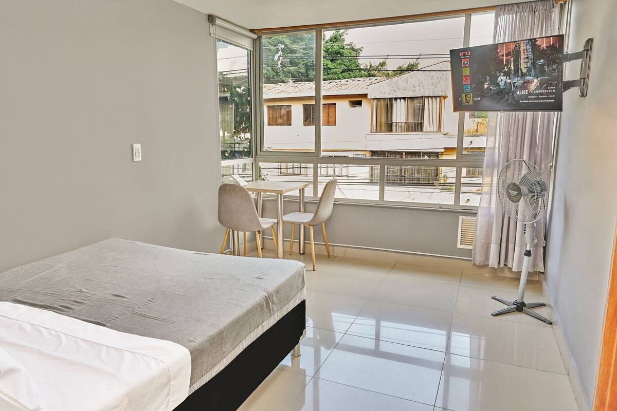 Een hotelkamer met een bed, een bureau, twee stoelen, een televisie en een raam met uitzicht op gebouwen.