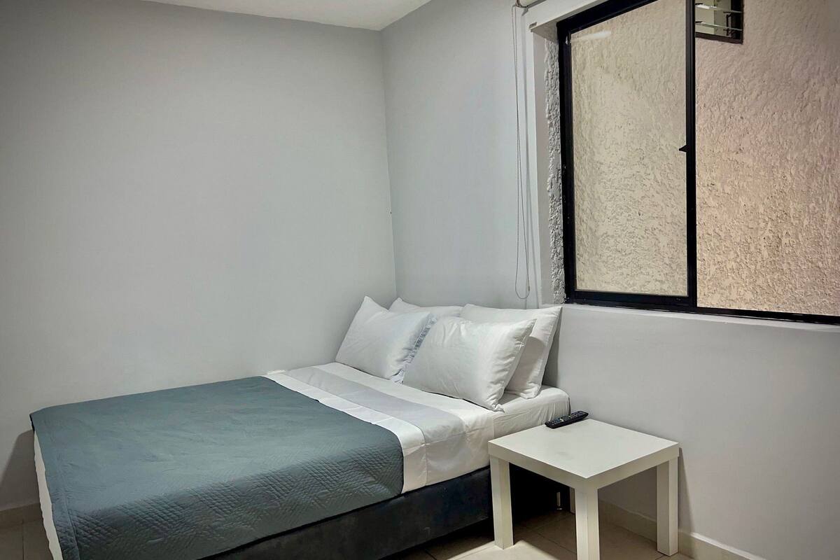 Een slaapkamer met een bed, nachtkastje, raam en een spiegel aan de muur.