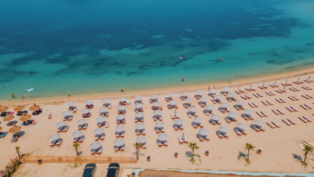 Vista aérea de una playa con filas de tumbonas y sombrillas, un mar azul cristalino y una montaña al fondo.